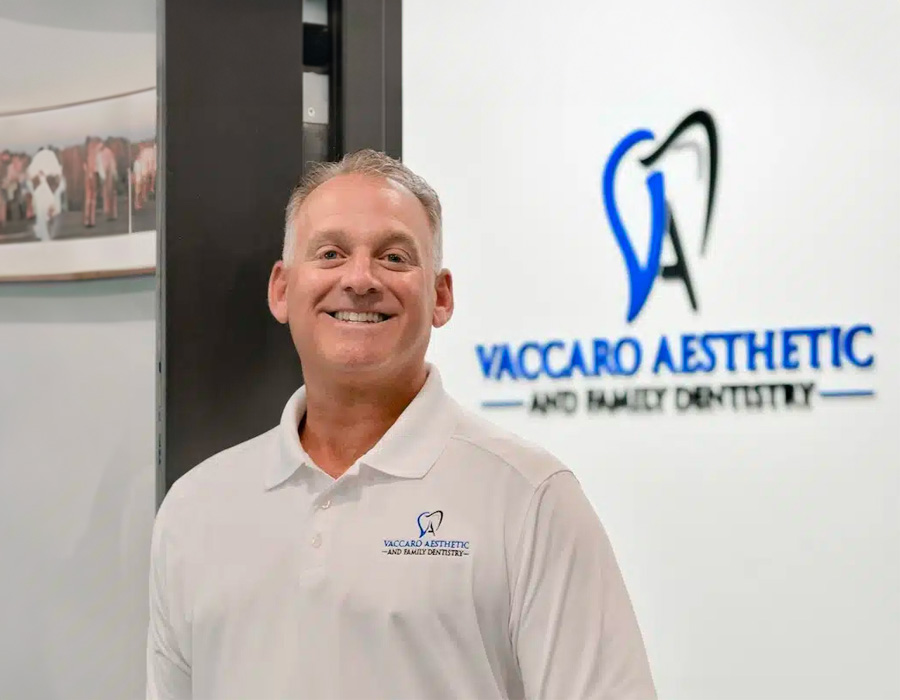 The image shows a man standing in front of a sign for Vaccaro Aesthetic Dentistry, smiling at the camera.