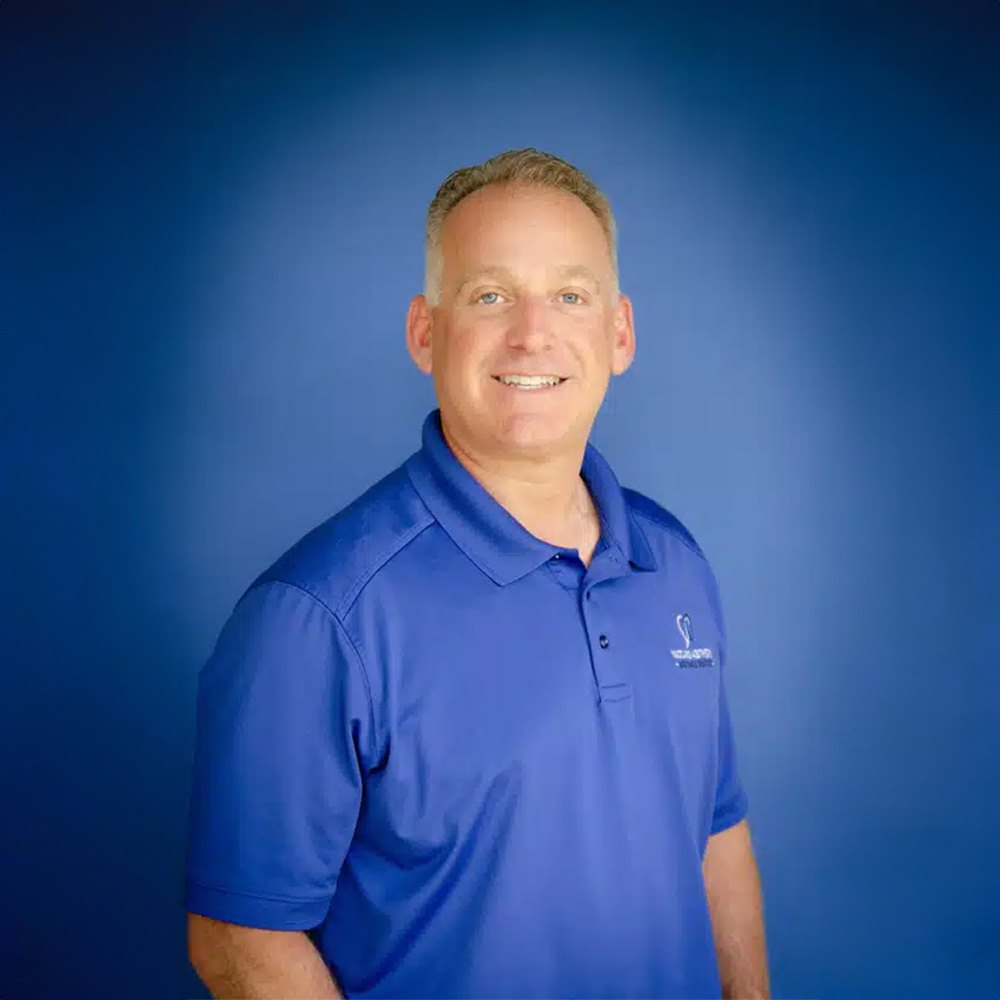 The image shows a man standing against a blue background, wearing a polo shirt and smiling at the camera.