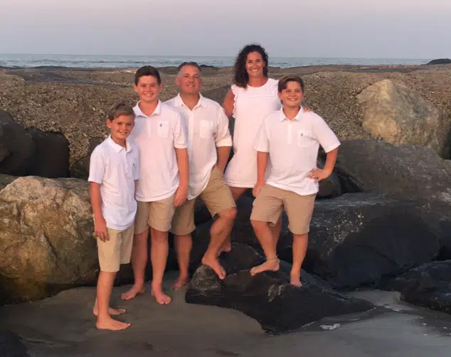 The image shows a group of five individuals posing on a rocky shoreline at sunset, with a body of water visible in the background. They are dressed in casual attire, including white shirts and khaki shorts, suggesting a relaxed and informal setting.