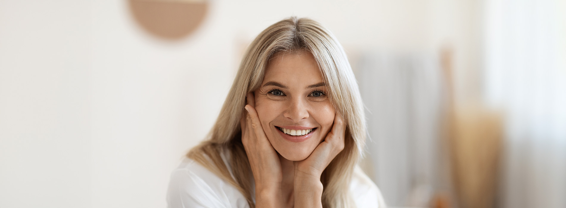 The image shows a woman with her hand on her face, smiling at the camera, set against a blurred background that suggests a domestic setting.
