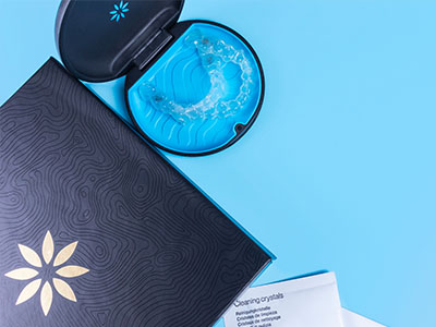 The image displays a closed package with a blue and black design next to an open package containing a product, which appears to be some sort of skincare item, possibly a mask, accompanied by a small cup and a spoon.