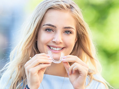 A smiling young woman holding up a clear retainer with her teeth.