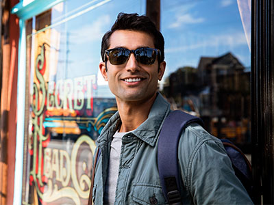 A man wearing sunglasses and a backpack stands outside a storefront with a sign visible through the window.