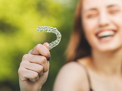 The image shows a person holding up a toothbrush with a smile on their face against a blurred background.