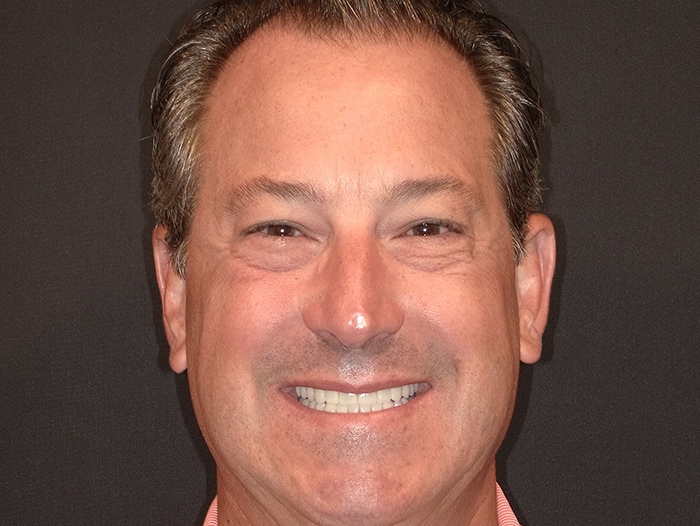 The image shows a man with a smile, wearing a pink shirt, against a dark background.