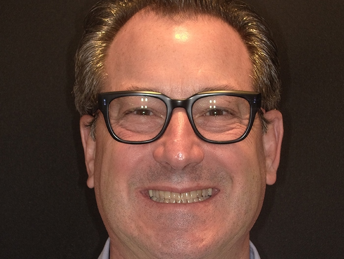 The image shows a man wearing glasses and a smile, with his head cropped from just below the hairline down to the chin.