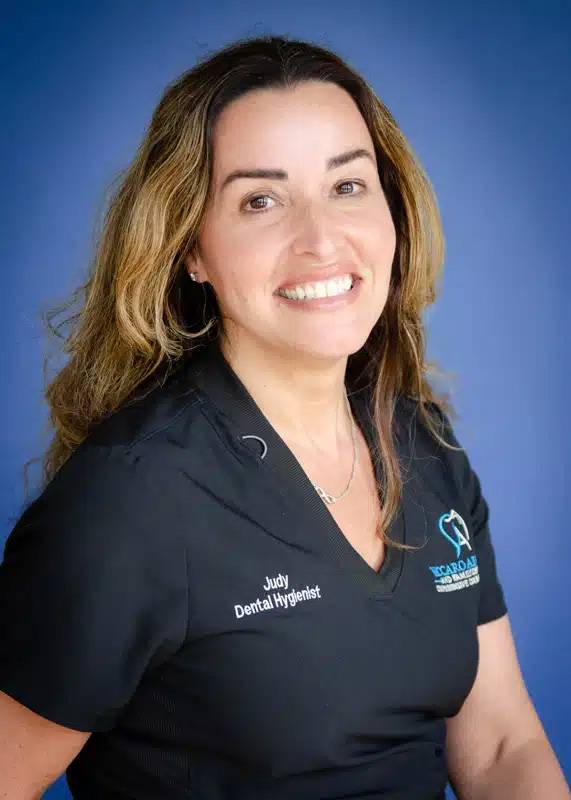 The image features a woman with dark hair smiling at the camera, wearing a black polo shirt with a name tag on the left side of her chest, posing against a blue background.