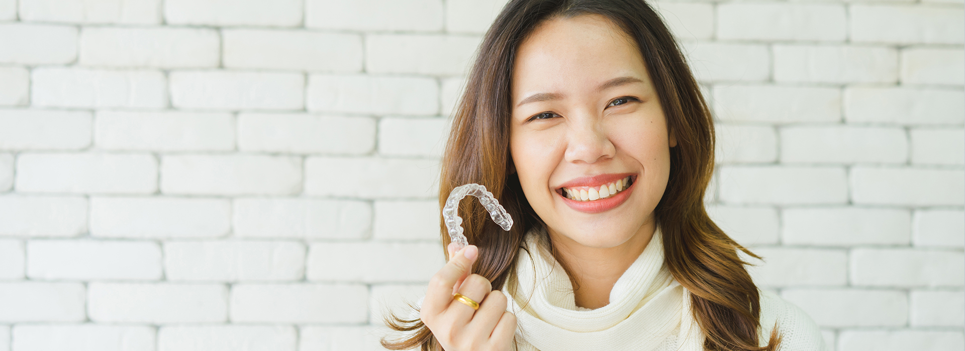The image shows a person holding up a toothbrush with a smile, against a white brick background.