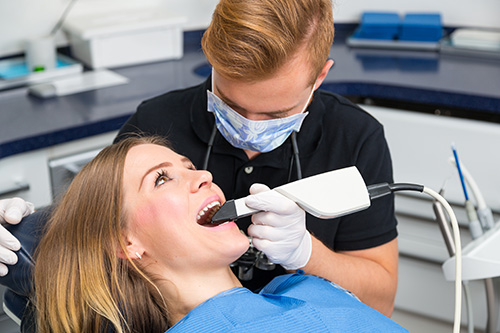 The image shows two people in a dental office setting one person is seated in a dental chair receiving dental treatment, while another person stands behind them, wearing a surgical mask and using a device that appears to be part of the dental equipment.