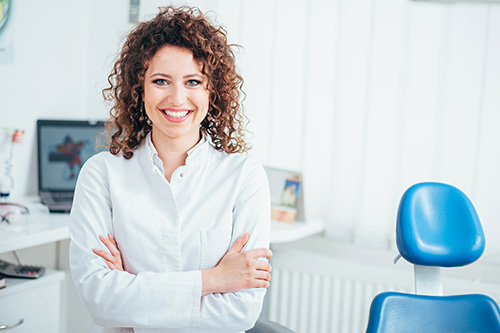 The image features a woman standing confidently in a dental office setting, smiling at the camera while leaning against a chair with her arms crossed.