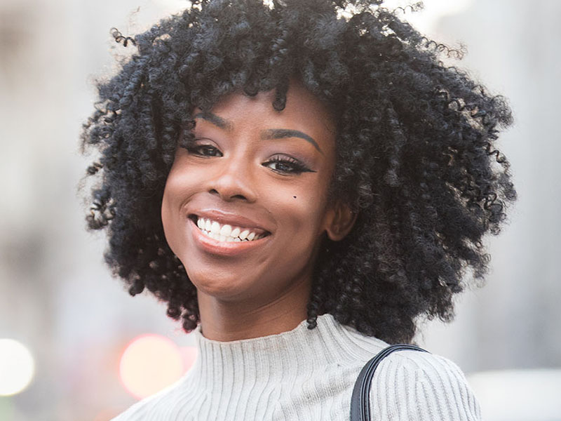 The image shows a person with curly hair smiling at the camera, wearing a dark top and a light-colored sweater, with a blurred background suggesting an urban setting.